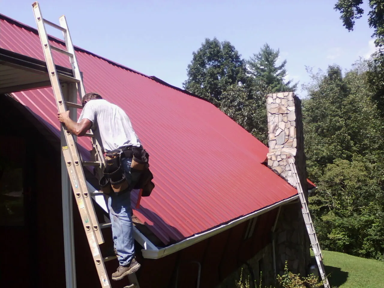 Roofer on a ladder beside a red metal roof and stone chimney — Soffit & Fascia Repair by Roofing Brothers in Twain Harte